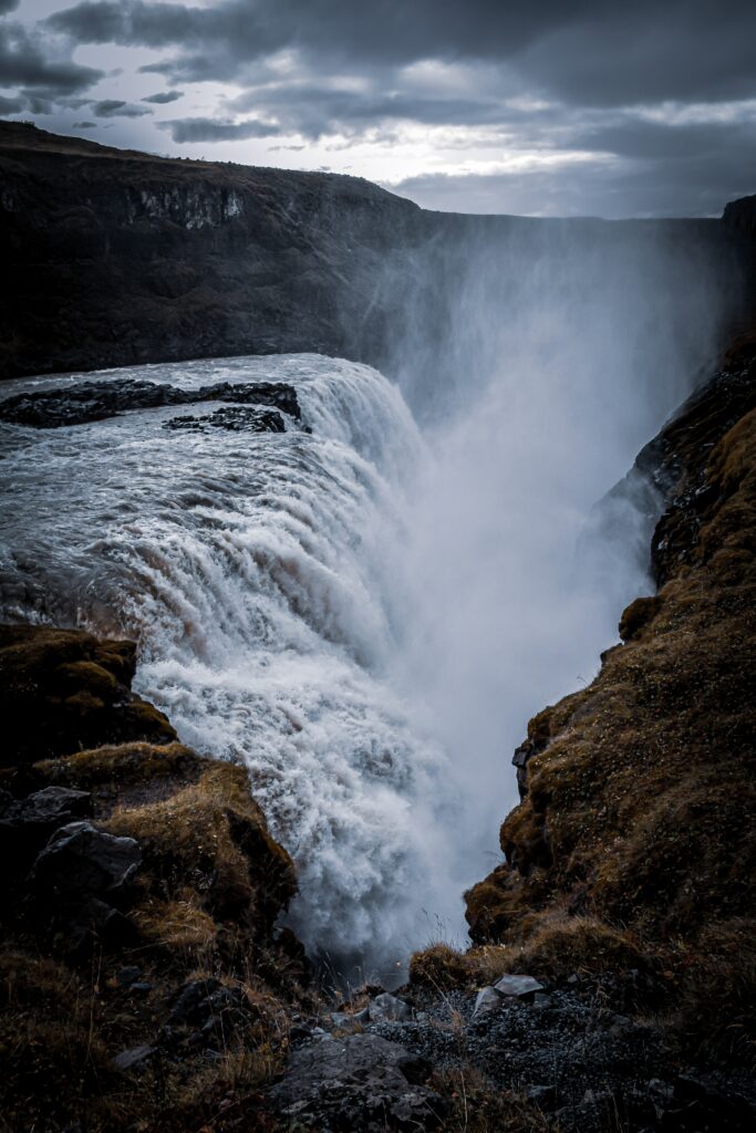 Dramatic waterfall cascading over rugged cliff into rocky gorge with misty spray and dark skies Wallpaper
