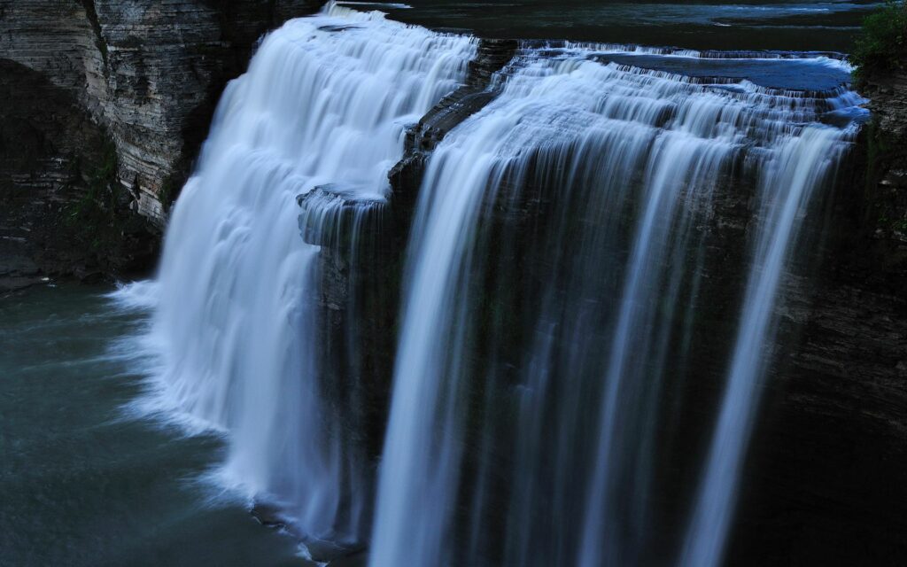 Dramatic waterfall cascading over rugged rocks with cool blue tones and shadowy natural landscape Wallpaper