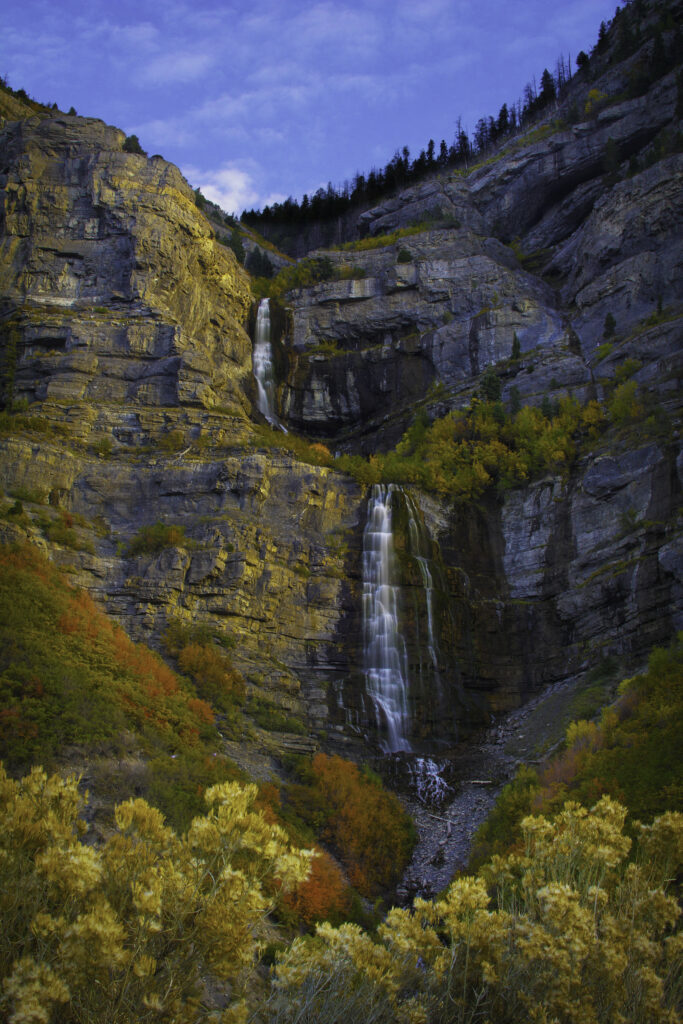 Waterfall cascading over rocks surrounded by colorful autumn trees and lush greenery under blue sky Wallpaper