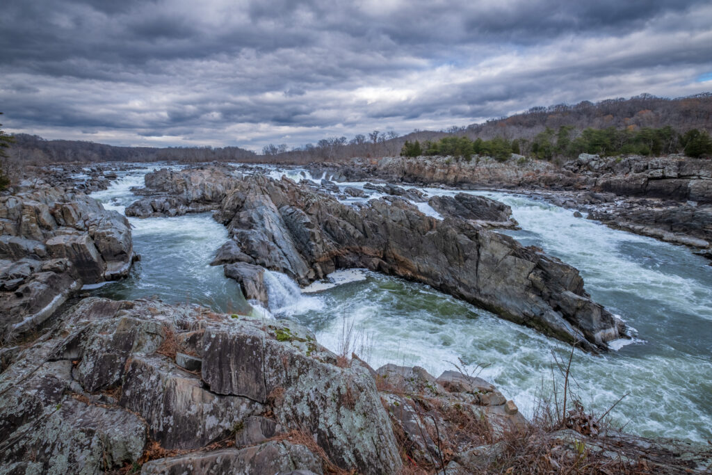 Dramatic river cascades over jagged stones with winter trees under moody cloudy sky wallpaper