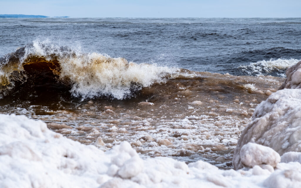 Dramatic wave crashes icy shore with snow covered rocks and dark churning ocean water wallpaper