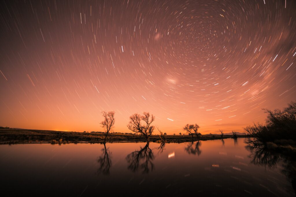 Starry night sky with swirling stars over silhouetted trees reflected in calm water at dusk Wallpaper