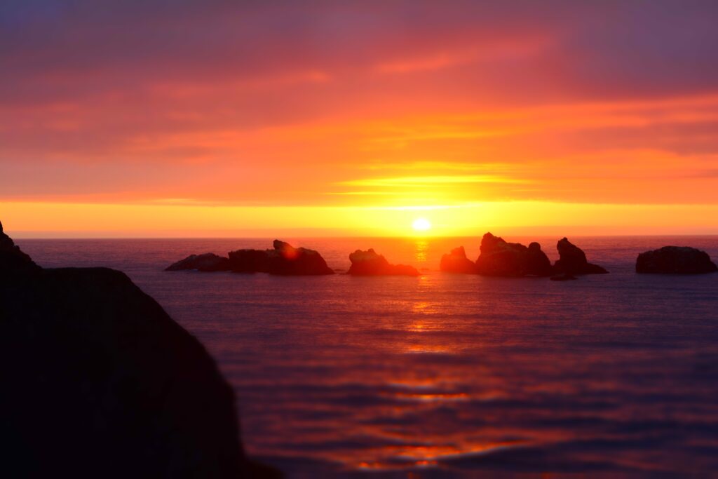Sunset over sea with glowing sun reflecting on water and jagged rocks in colorful sky Wallpaper