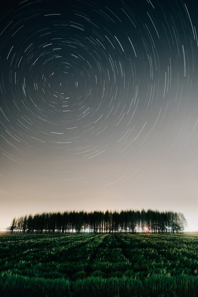 Star trails swirl over a tranquil field bordered by tall trees in a mesmerizing night sky wallpaper