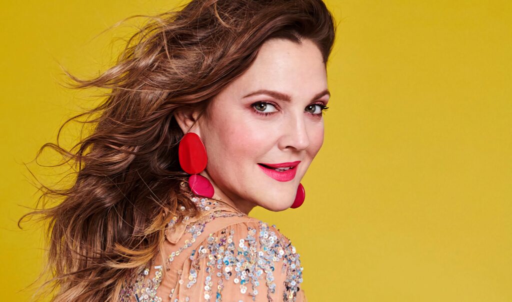 Drew Barrymore confident in sequined top and red earrings on vibrant yellow background wallpaper