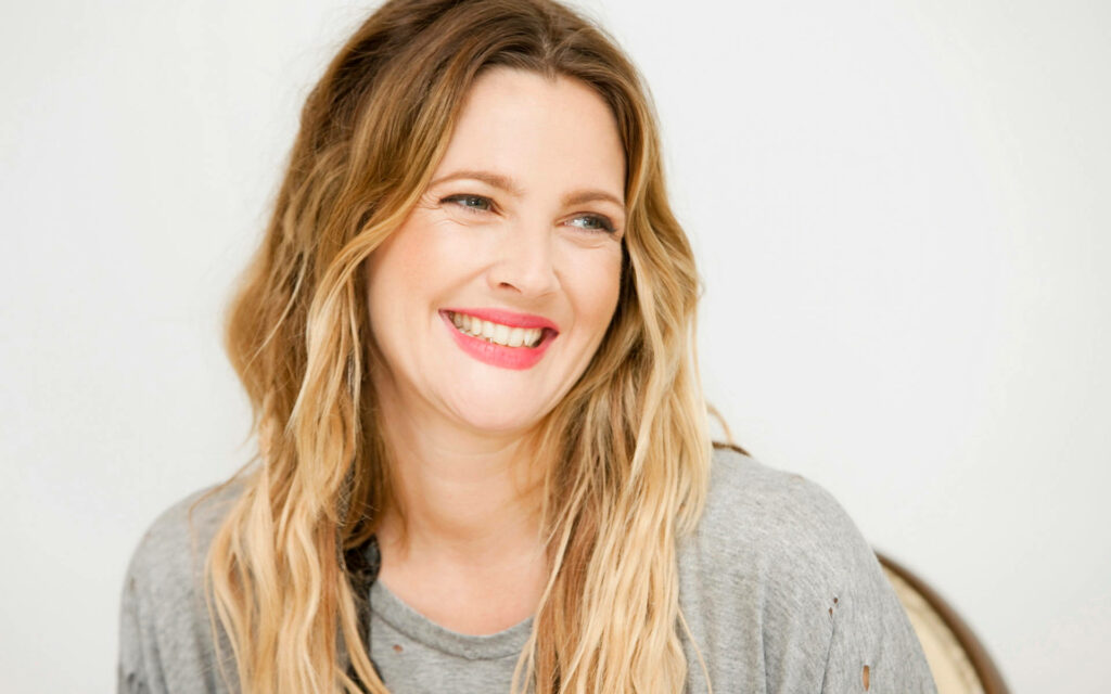 Drew Barrymore smiling warmly with wavy hair and gray top on bright soft wallpaper background