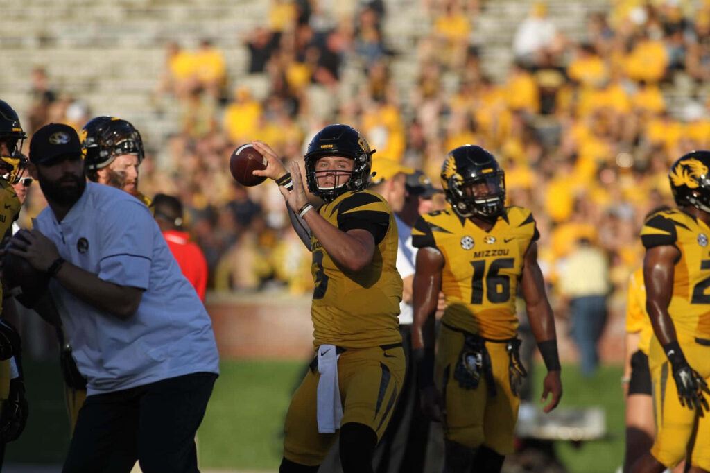 Drew Lock Mizzou quarterback mid-throw in yellow black uniform on sunny game day with crowd Wallpaper