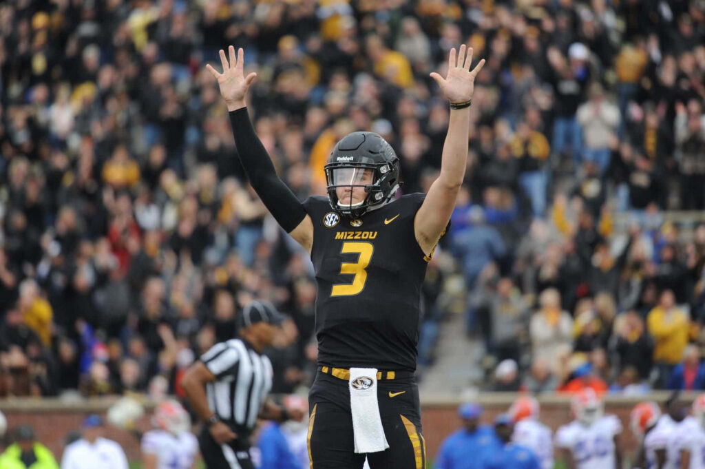 Drew Lock celebrating a Missouri touchdown in black and gold uniform with stadium crowd background Wallpaper