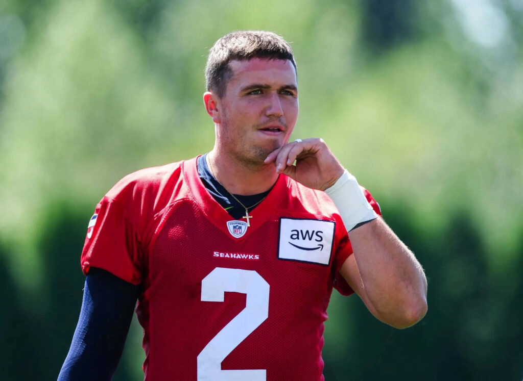 Drew Lock in red Seahawks training jersey practicing football outdoors with blurred green background Wallpaper