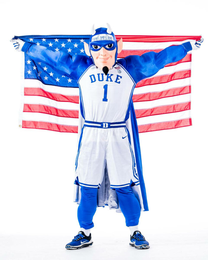 Duke Blue Devils mascot in blue white costume with cape poses before vibrant American flag wallpaper