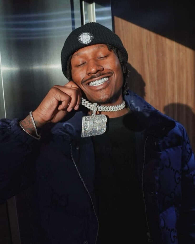 Duke Dennis in elevator wearing black beanie patterned jacket and sparkling chain smiling brightly wallpaper