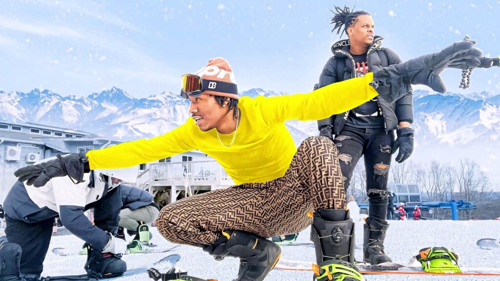 Duke Dennis in stylish winter gear posing dynamically on snowy mountain with vibrant alpine backdrop Wallpaper