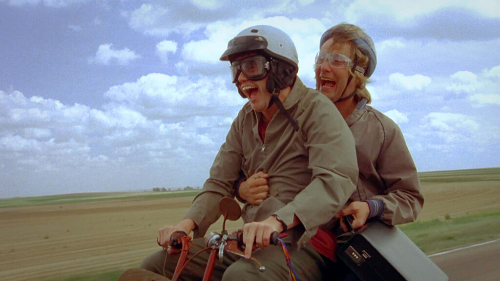 Jim Carrey and Jeff Daniels as Dumb and Dumber riding motorbike in colorful countryside scene Wallpaper