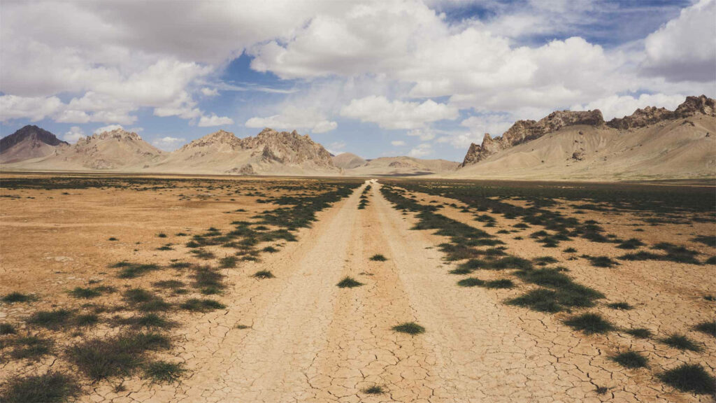 Dusty pathway across cracked Afghan desert earth with rugged mountains and cloudy sky background Wallpaper