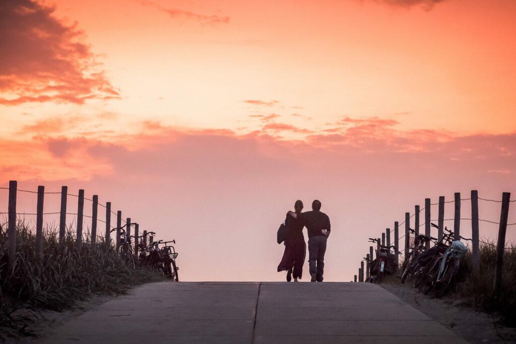 Couple walking arm in arm on Dutch dune path at sunset with peach sky and bicycles wallpaper