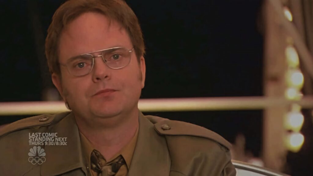 Dwight Schrute in khaki jacket and glasses serious expression dim lit warm blurred background wallpaper