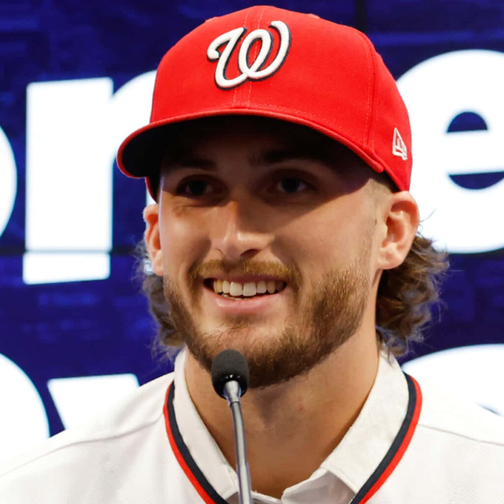 Dylan Crews smiling in red Nationals cap and white jersey at vibrant sports press conference background Wallpaper