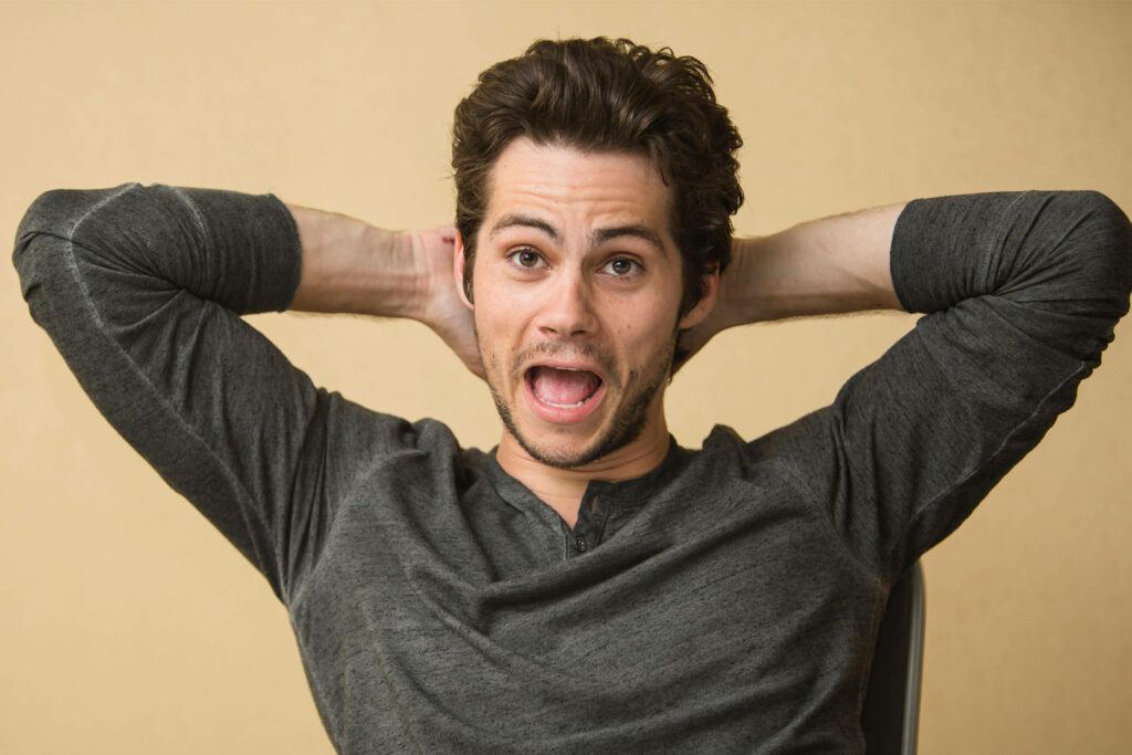 Dylan OBrien in candid shocked expression hands behind head neutral beige background wallpaper
