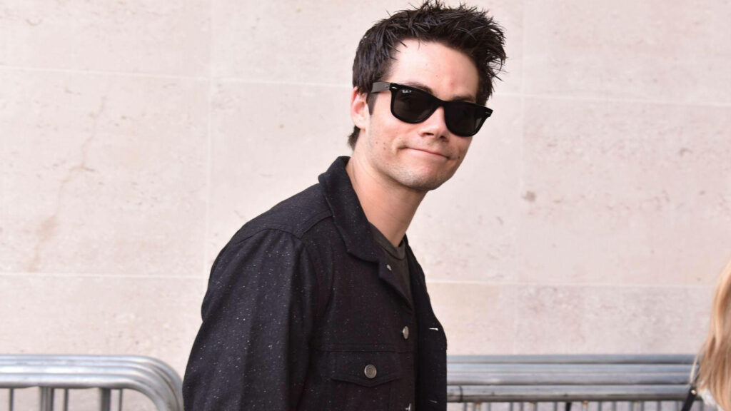 Dylan O'Brien in black jacket and sunglasses against stone wall cool casual style wallpaper background