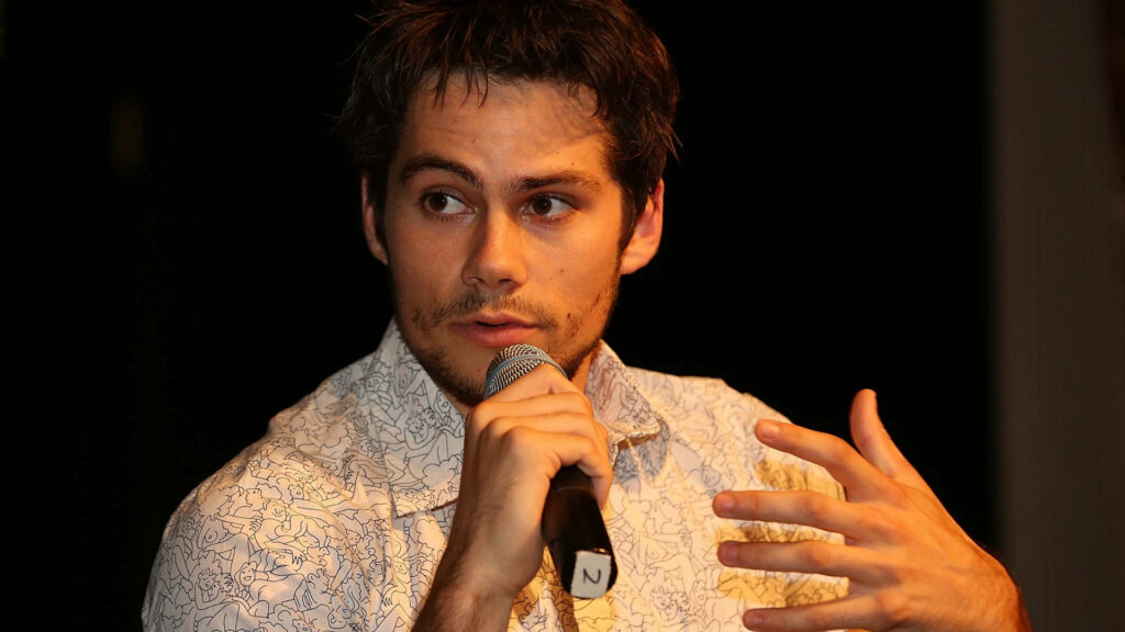 Dylan O'Brien speaking at press conference holding microphone warm lighting patterned shirt Wallpaper
