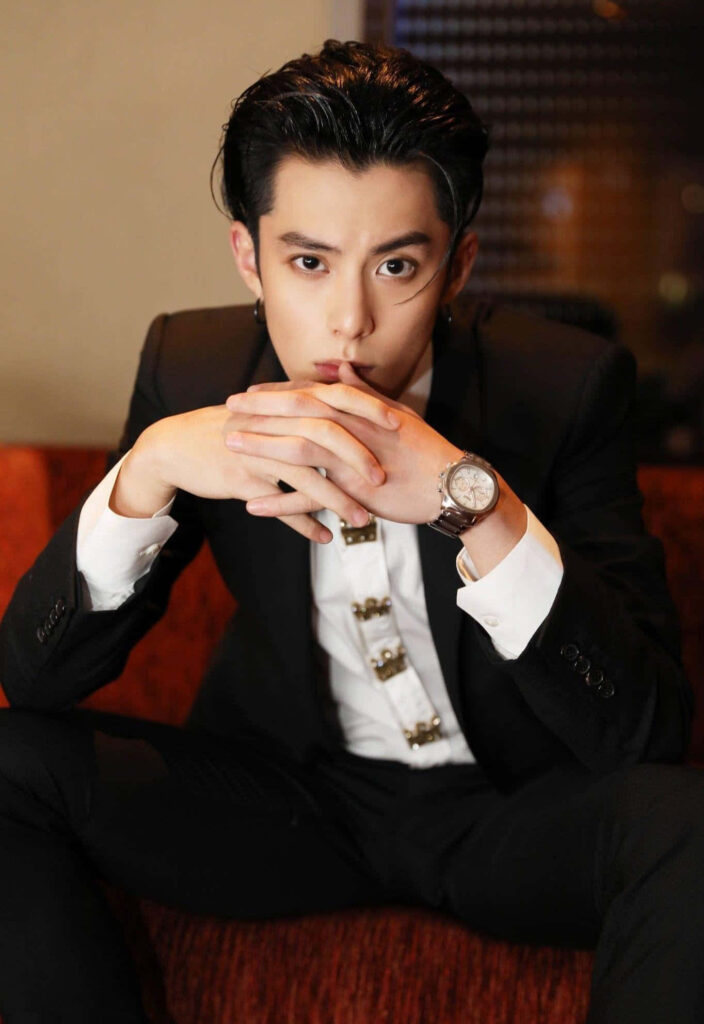 Dylan Wang in sharp black suit with metallic details sitting on red couch in warm soft light Wallpaper