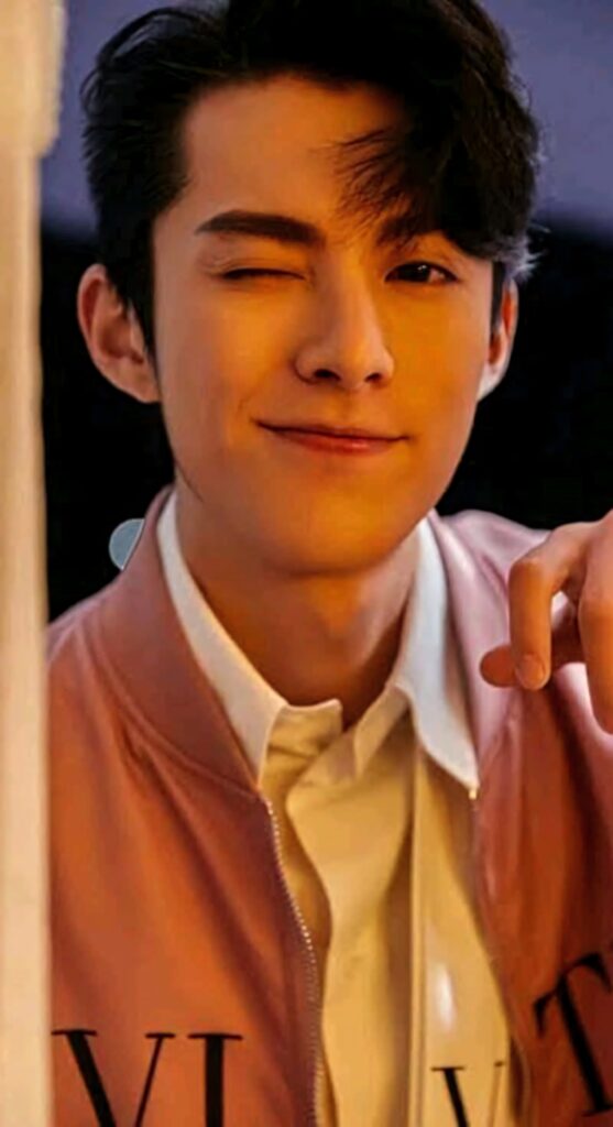 Dylan Wang winking wearing pink jacket and white shirt with warm lighting stylish wallpaper background