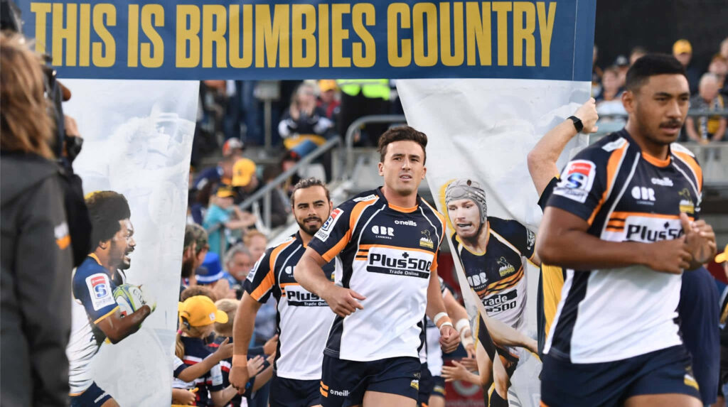 Dynamic ACT Brumbies rugby players entering field with bold navy gold jerseys and team banner Wallpaper