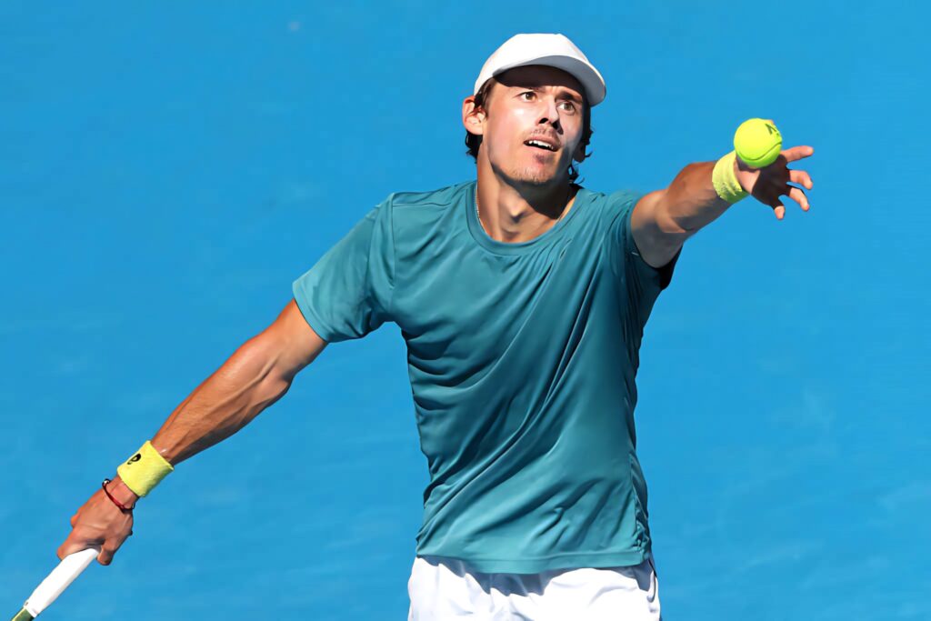 Alex de Minaur serving powerfully on tennis court with vibrant blue background wallpaper