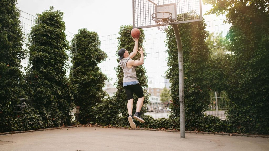 Basketball player in grey hoodie jumps to score on outdoor court with ivy and sunlight background Wallpaper