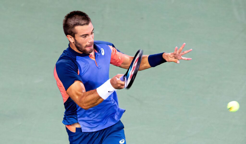 Borna Coric mid-action tennis shot wearing blue and orange sportswear high-energy court photo Wallpaper