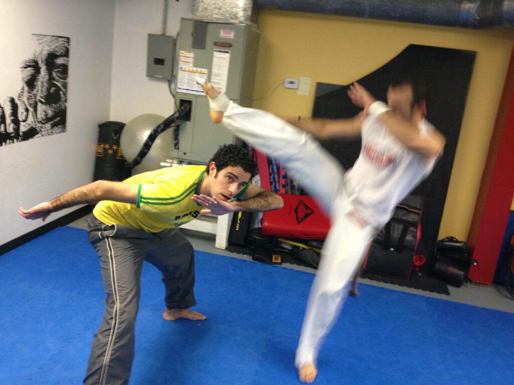Two athletes practicing Capoeira in gym one performs high kick other skillfully dodges dynamic motion Wallpaper