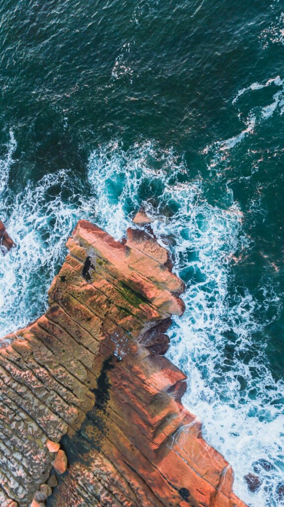 Aerial view of vibrant turquoise waves crashing rugged layered rocks on dynamic coastal shore Wallpaper