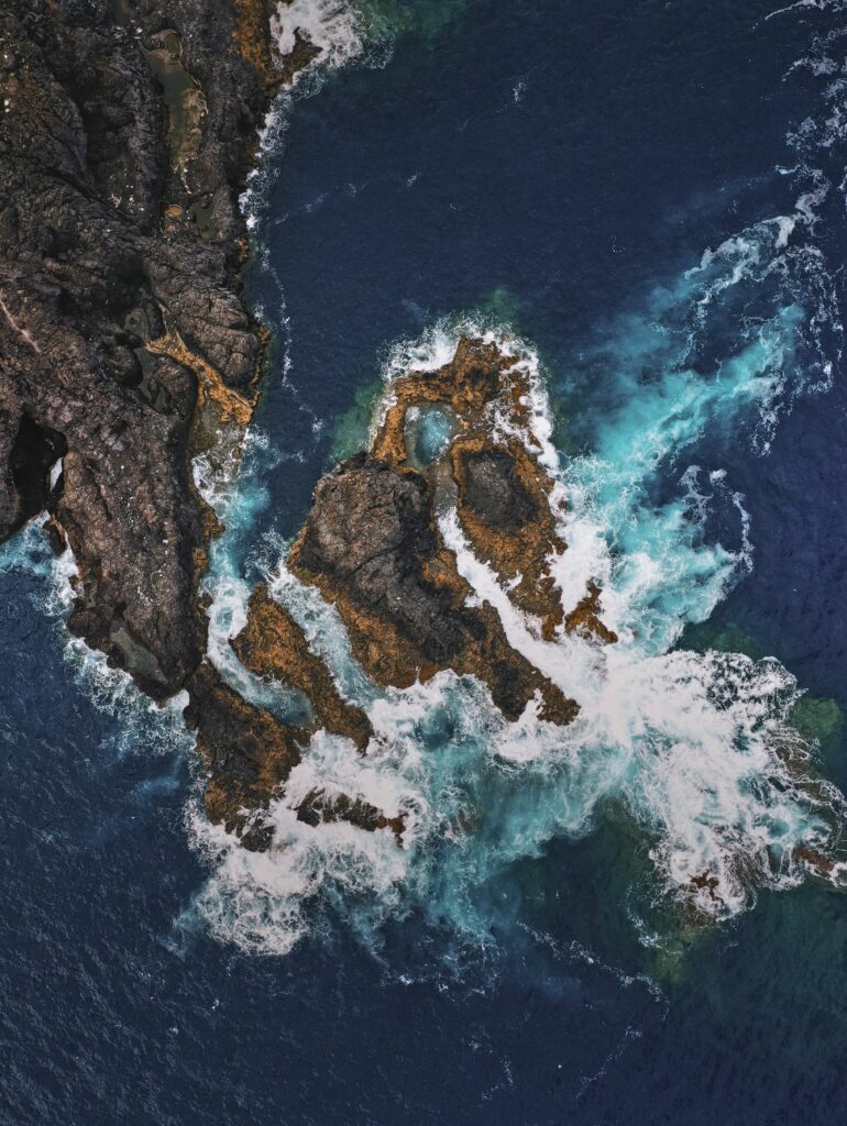 Aerial view of deep blue sea crashing on rugged coastal rocks with foamy waves shore wallpaper