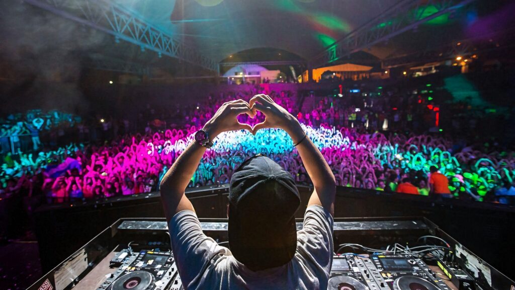 DJ performing on vibrant stage with colorful lights and hands forming heart to cheering crowd Wallpaper