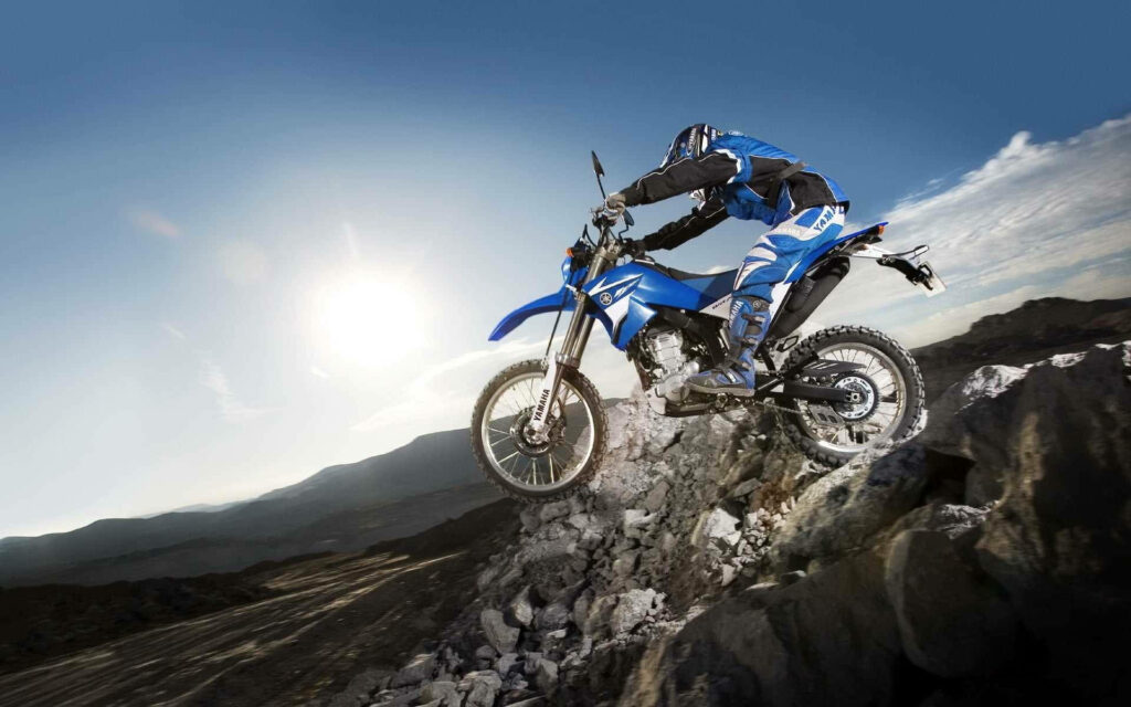 Dirt bike rider in blue gear climbing rocky hill under dramatic sunlit sky off-road adventure Wallpaper