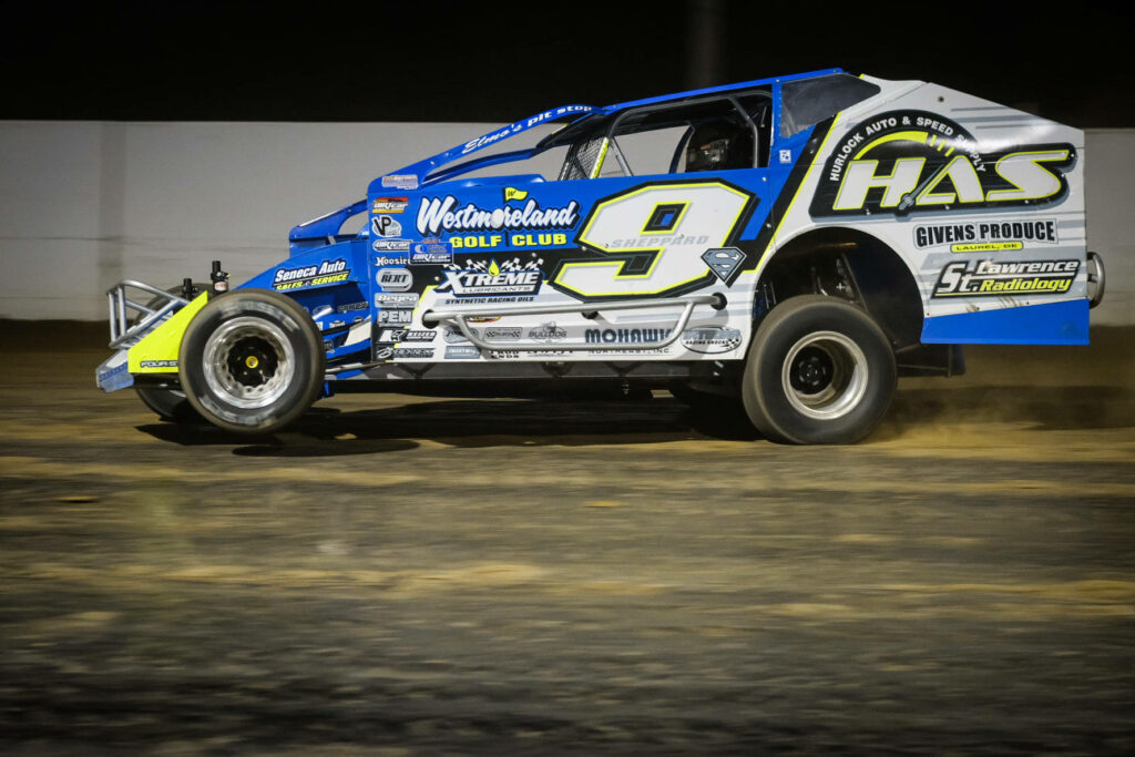 Dirt car number 9 with blue yellow and white graphics racing on illuminated dirt track at night Wallpaper