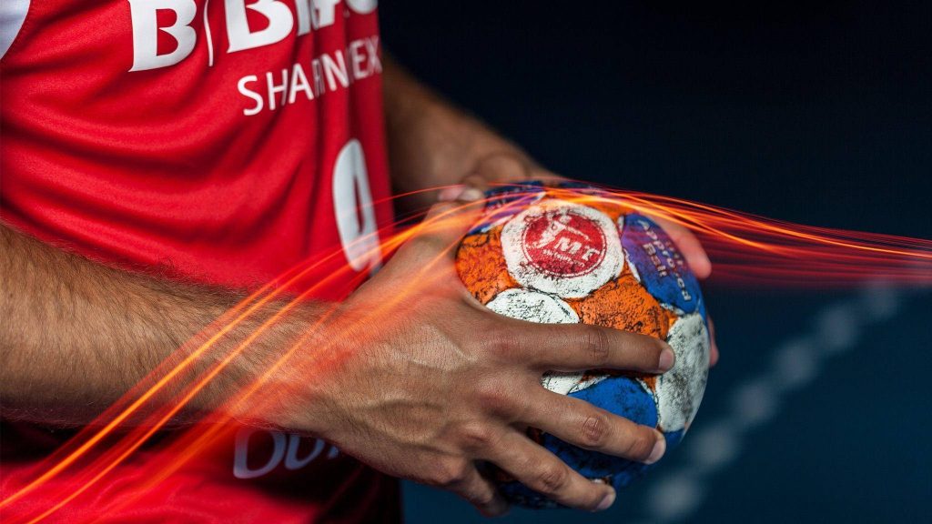 Handball player in red jersey gripping vibrant orange white blue ball with dynamic light trails Wallpaper