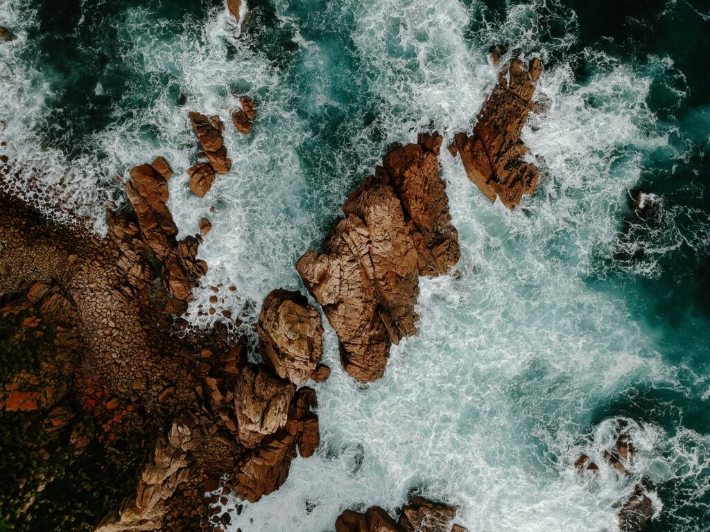 Aerial view of ocean waves crashing rugged brown rocks with foamy white water coastal wallpaper