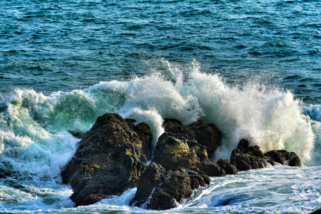 Vivid blue ocean waves crash on rugged dark rocks sending white foam and splashes into air Wallpaper