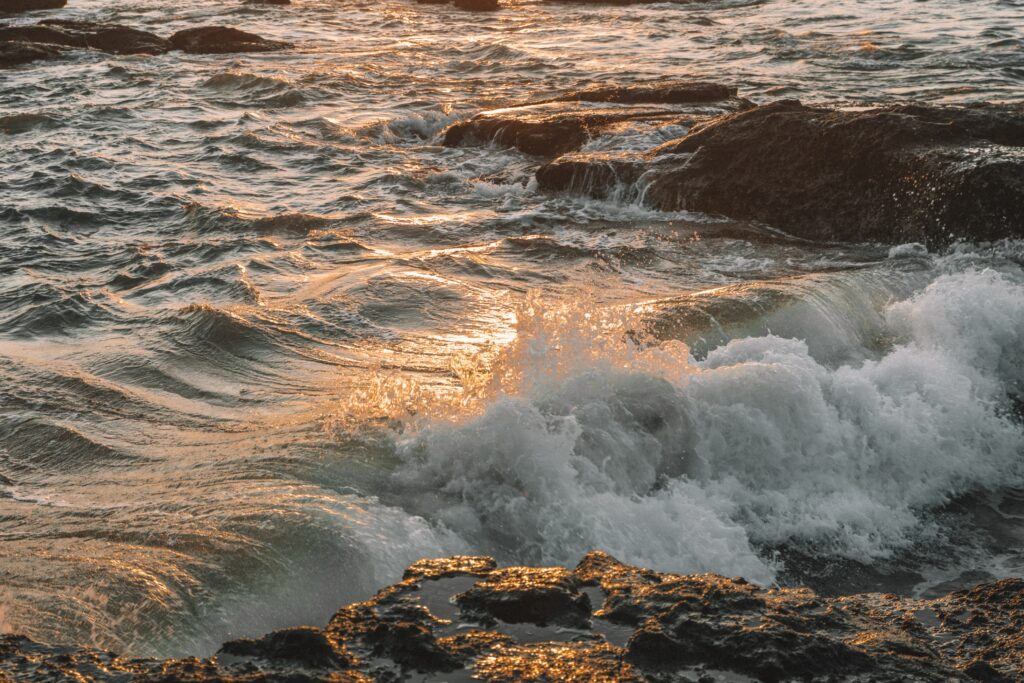 Ocean waves crashing on rugged rocks with sunlight reflections creating a serene coastal scene Wallpaper