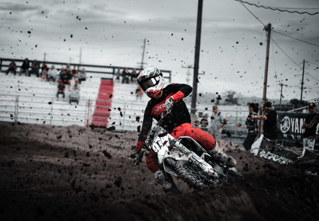 Dirt bike rider in red and black gear speeds through muddy motocross track with crowd background Wallpaper