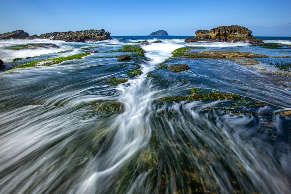 Vibrant sea whirlpool over mossy stones with blue sky and rocky outcrops nature wallpaper background