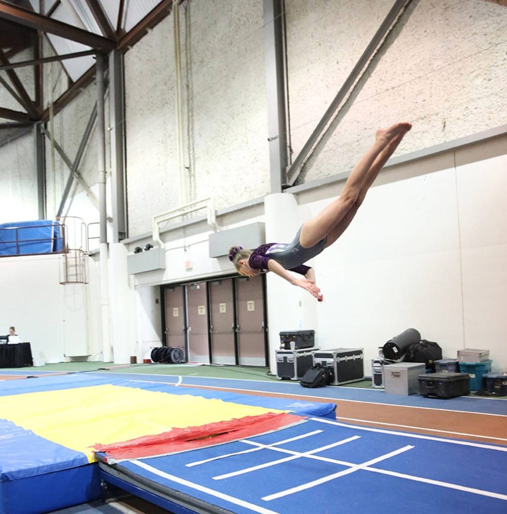 Gymnast soaring mid-air in trampoline and tumbling gym above blue mats high-energy sports background Wallpaper