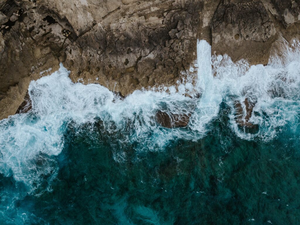 Aerial view of turquoise ocean waves crashing against rugged rocky shore with white foam Wallpaper