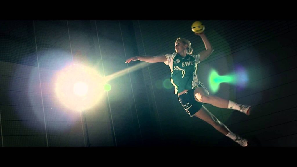 Dynamic female handball player in green jersey jumping to strike yellow ball on dark court background Wallpaper