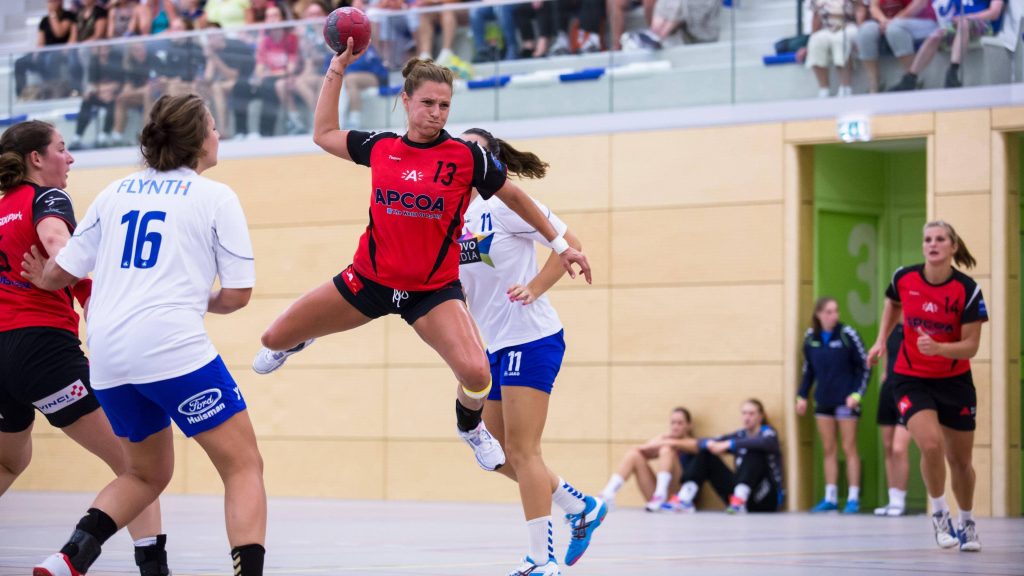 Female handball player in red black jersey jumping to shoot during intense indoor match action Wallpaper