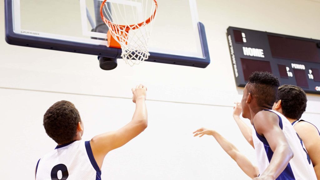 Three young basketball players in white navy jerseys leap toward hoop indoor court scoreboard Wallpaper