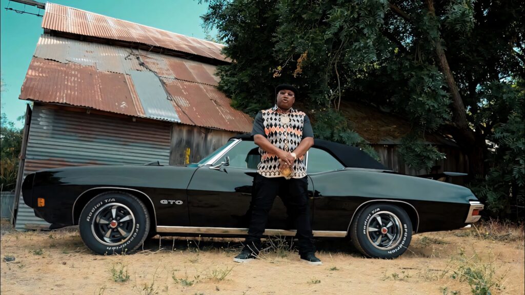 E40 posing by classic black GTO convertible near rustic shed with greenery hip hop urban style Wallpaper
