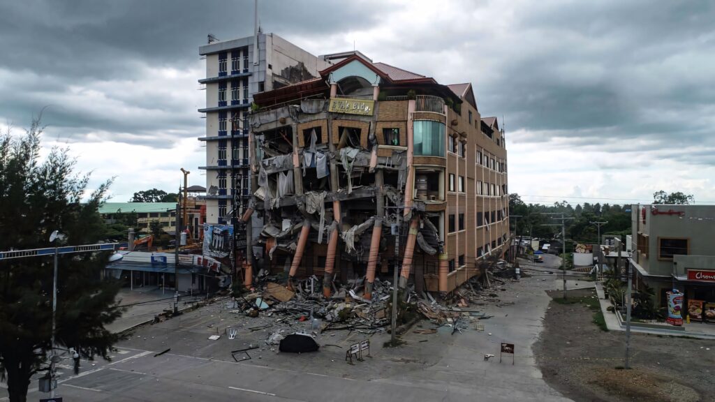 Severely damaged building after earthquake with collapsed columns debris and cloudy sky background Wallpaper
