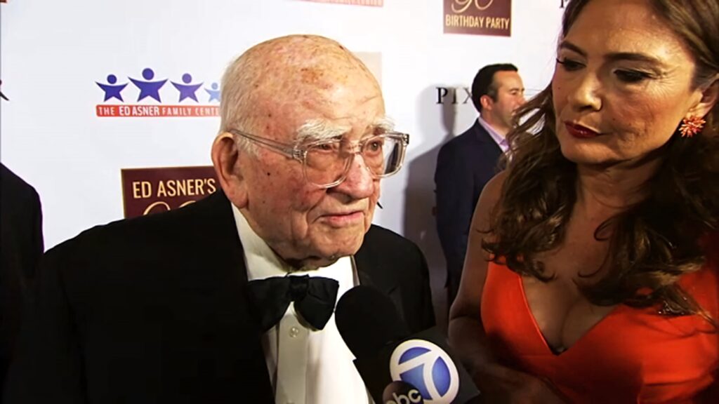 Ed Asner in black tuxedo interviewed by Navah Paskowitz at lively 90th birthday party celebration Wallpaper